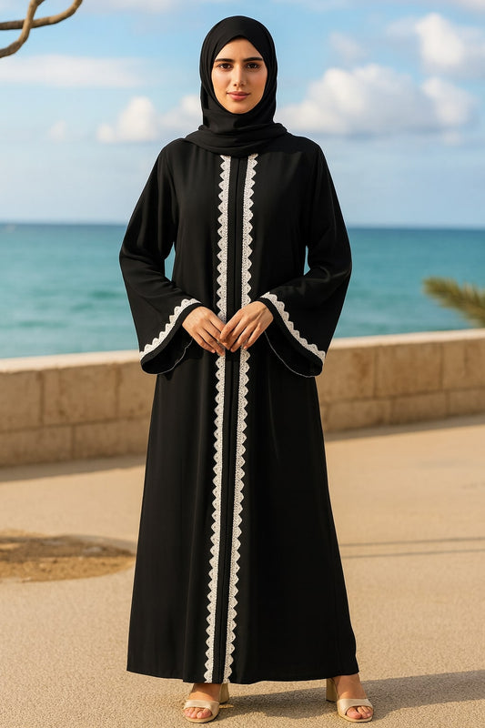 Woman wearing a black abaya with white trim standing by the ocean.