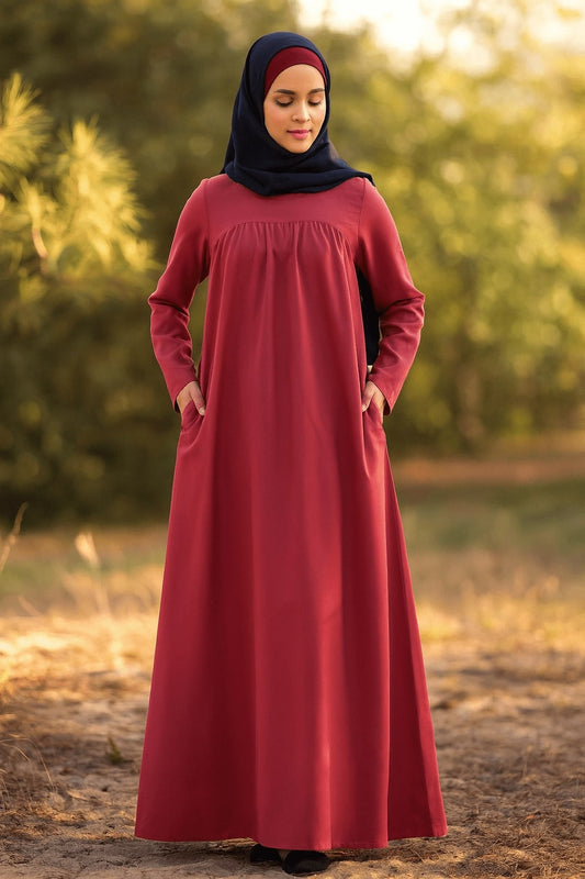 Woman wearing a long red dress and black hijab standing in a natural setting with trees and grass.