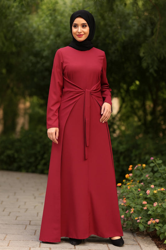 Woman wearing a red dress with a black hijab standing outdoors with greenery and flowers in the background.