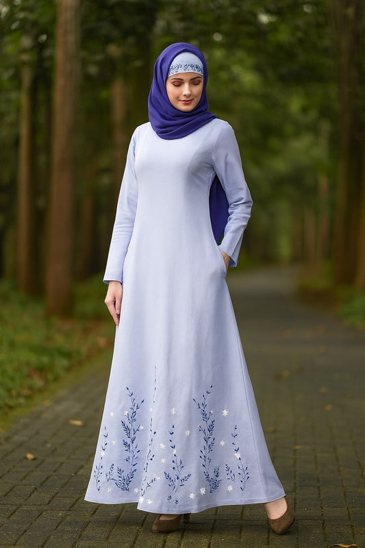 Woman in a long white dress with blue embroidery and a purple hijab standing in a forest.