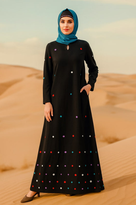 Woman in a black dress with colorful polka dots and a blue hijab standing in desert sand dunes.