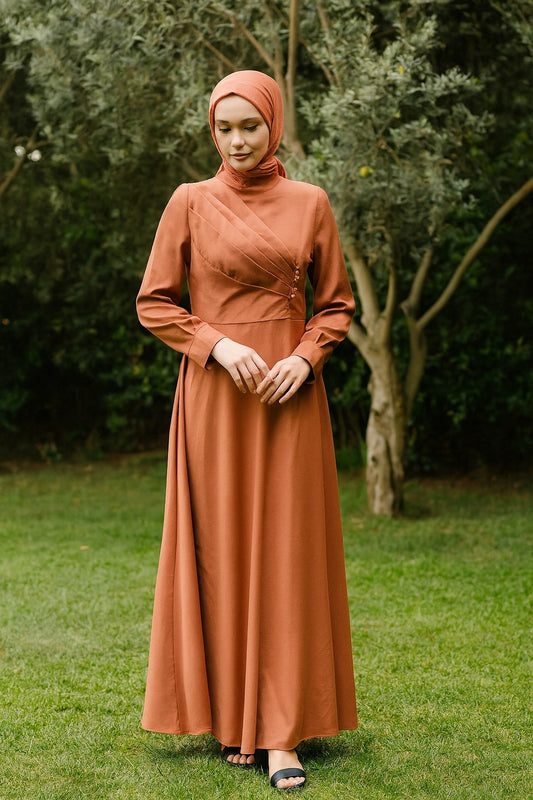 Woman wearing a long brown dress and hijab standing in a grassy area with trees.