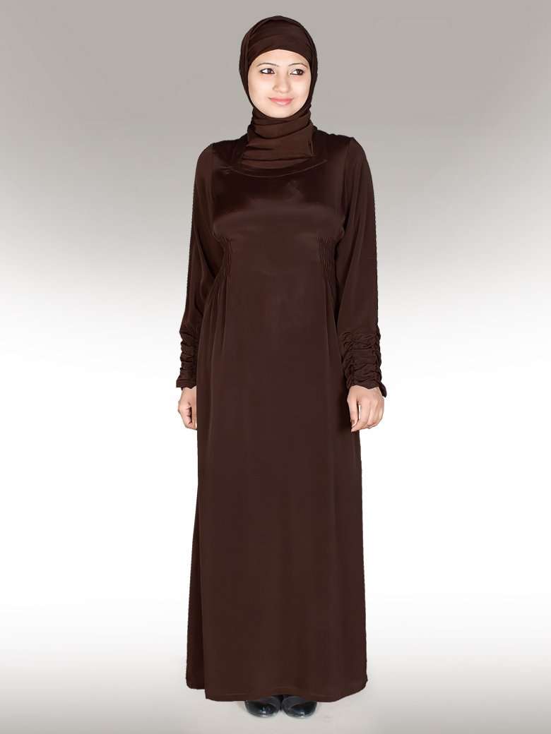 A woman wearing a brown abaya with a round neckline, long sleeves, and a flared bottom design. The abaya features side gathers on the waist and has utility pockets on the sides.