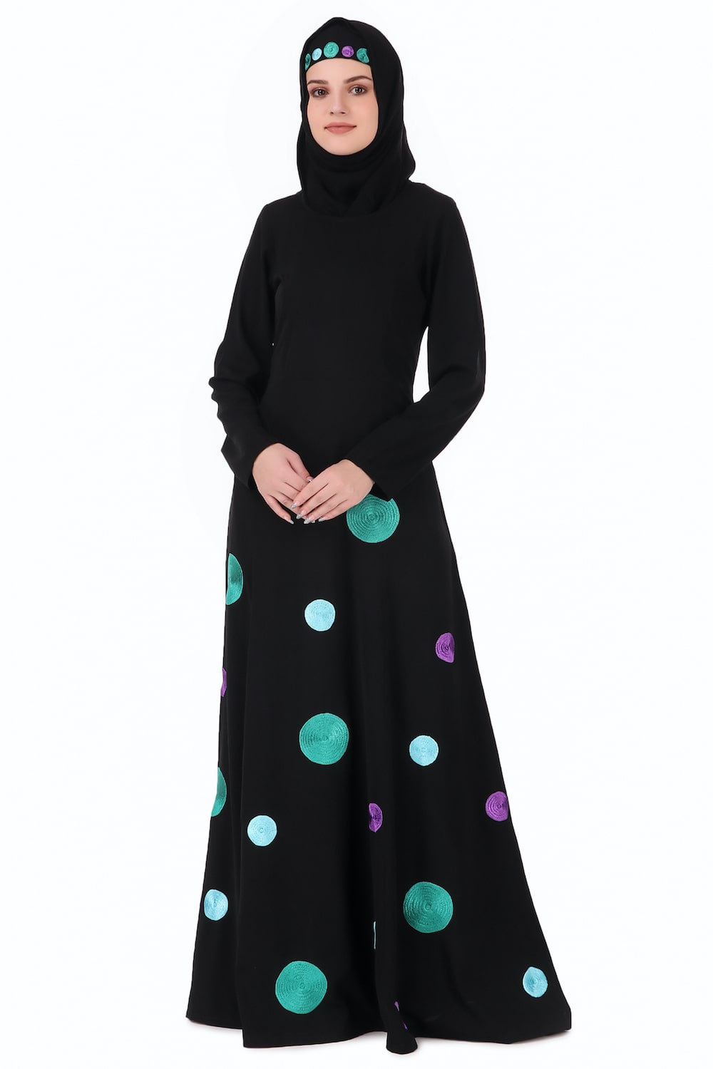 A woman wearing a black Nida abaya with round neckline and straight sleeves. The abaya features a front panel with shiny big and small polka dots embroidered below the waist line, and it has utility pockets on the sides.