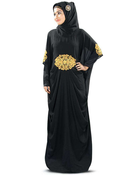 A woman wearing a black Kaftan with gold embroidery and a square or rectangle hijab. The Kaftan has a round neckline, pleats at the waistline, and matching embroidery at the sleeve ends.