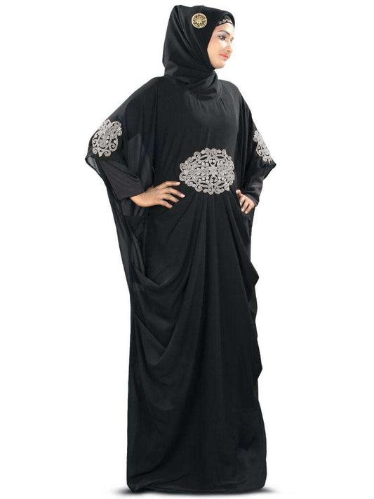 A woman wearing a black Kaftan with silver embroidery and a round neckline, standing against a white background.