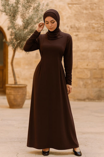 Woman wearing a brown dress and hijab standing in front of a stone wall.