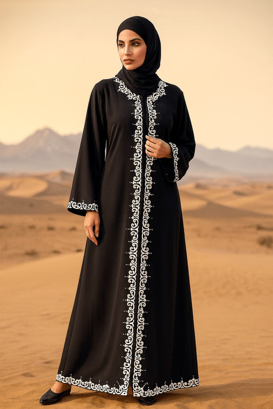 Woman in a black abaya with white embroidery standing in a desert landscape.