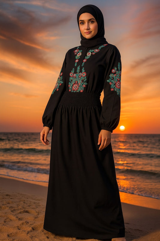 Woman in a black dress with floral embroidery standing on a beach at sunset.