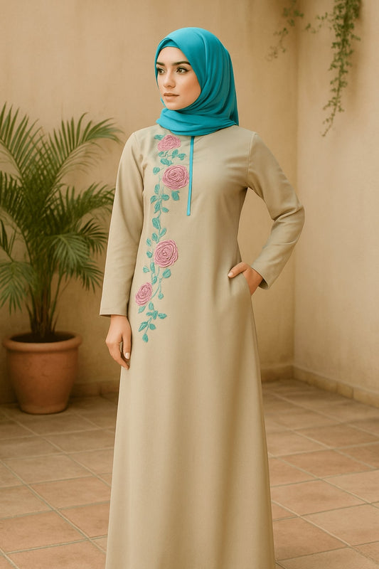 Woman wearing a beige dress with floral embroidery and a blue hijab standing in a room with a plant.