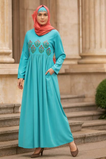 Woman wearing a turquoise dress with floral embroidery and a red hijab standing on steps.