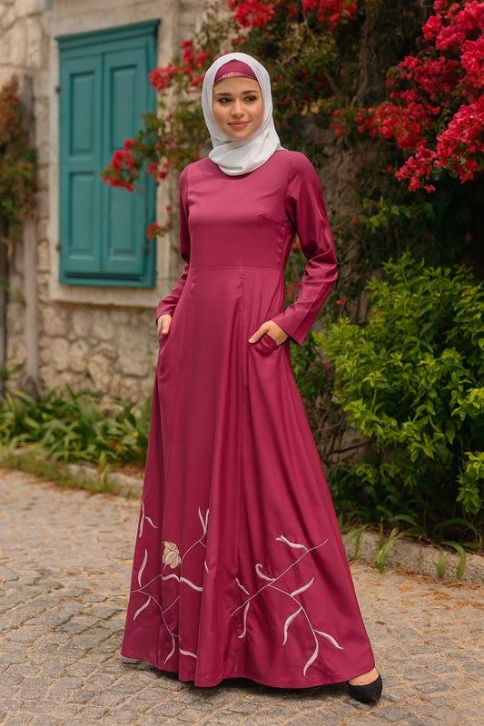 Woman in a burgundy dress with white embroidery standing outdoors near a stone building and red flowers.