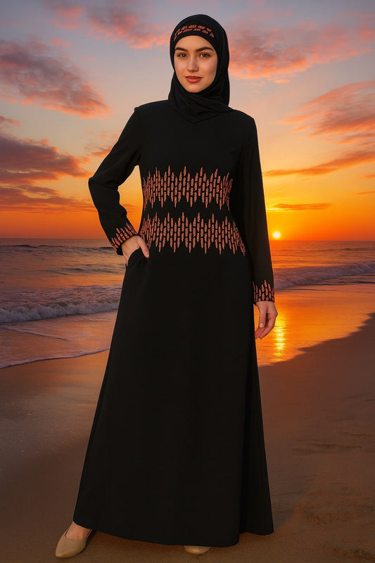 Woman in a black abaya with orange patterns standing on a beach at sunset.