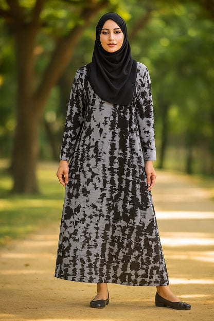 Woman wearing a patterned dress and black hijab standing outdoors with greenery in the background